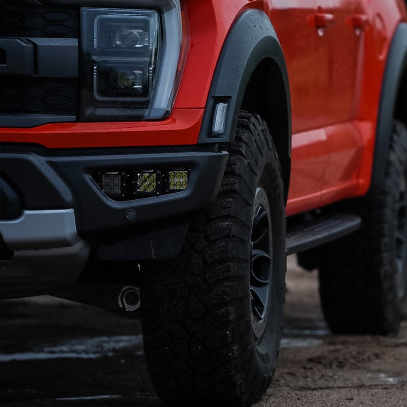 Rigid D-Series PRO Pods Pair installed on a Ford F-150 Raptor: three D-Series pods set in the lower bumper fog pocket of a red Ford F-150 Raptor beside its front tire