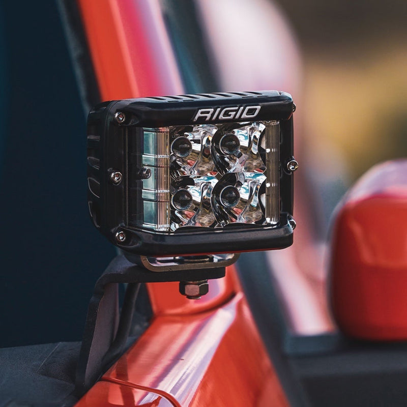 Rigid D-SS PRO Pods Pair installed on a vehicle: A black D-SS PRO side-shooter pod sits on an A-pillar bracket beside the mirror of a red vehicle