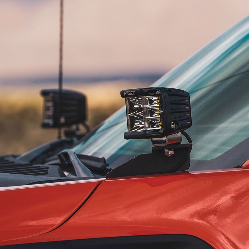 Rigid D-SS PRO Pods Pair installed on a vehicle: two D-SS PRO pods sit on A-pillar brackets of a red truck against a sunset sky