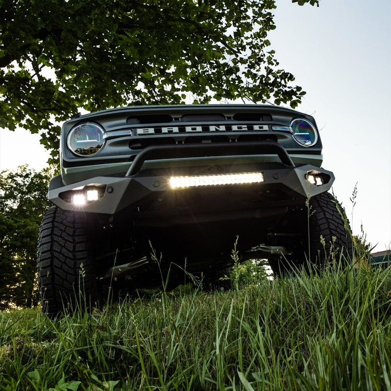 Rigid E-Series PRO 20" installed on a Ford Bronco: A green Ford Bronco with BRONCO grille lettering runs a lit LED light bar and two fog pods in its front bumper, seen from low in the grass