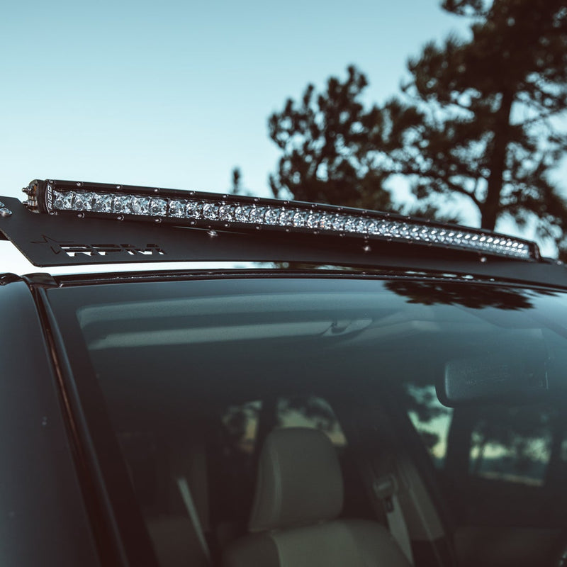 Rigid RDS SR-Series PRO 40" Spot installed on a vehicle: low-angle view of a curved single-row LED light bar mounted on a roof rack above a windshield, with pine trees and sky behind