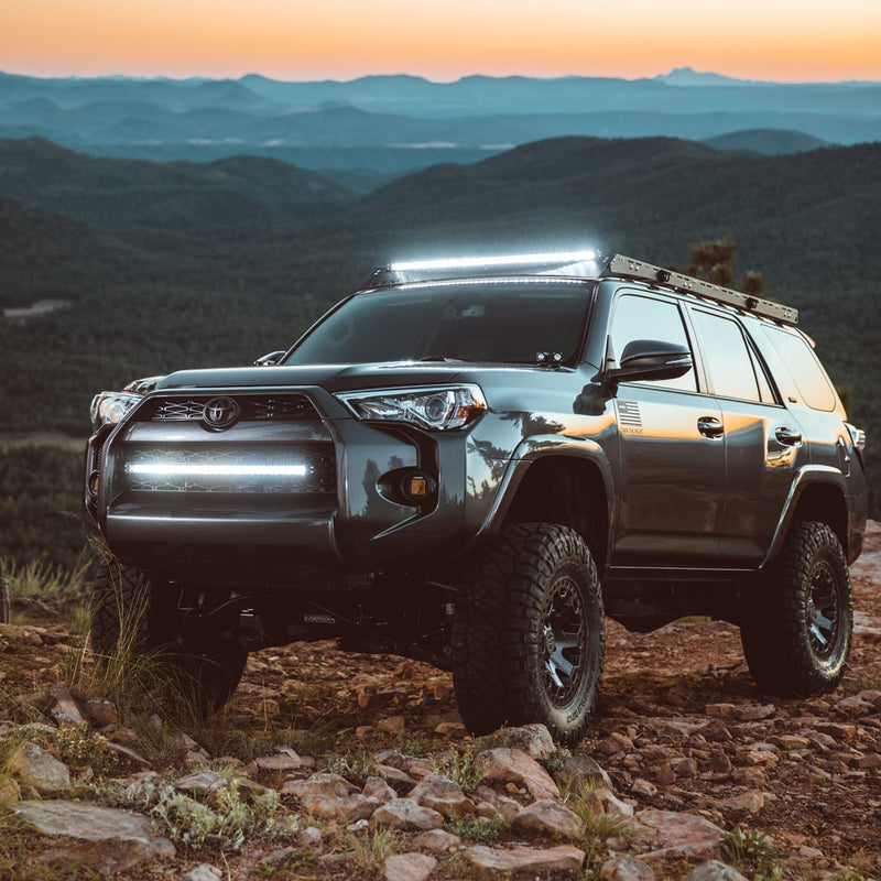 Rigid RDS SR-Series PRO 50" Spot installed on a Toyota 4Runner: A gray Toyota 4Runner on a rocky mountain ridge at sunset with a curved light bar lit on the roof and a bar lit in the lower grille