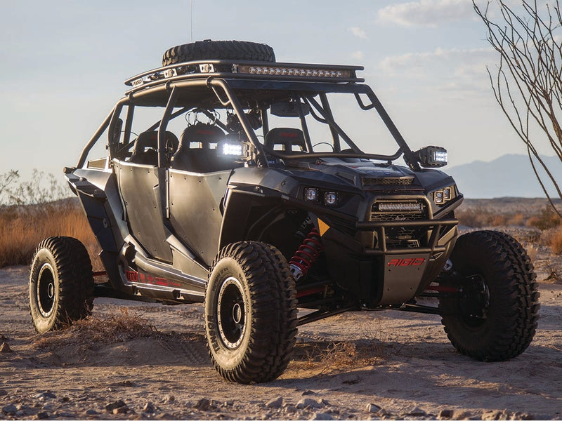 Rigid Reflect Side Mirror Lights Pair installed on a Polaris RZR XP 4: A four-seat side-by-side in the desert with both Reflect mirror lights glowing, a roof light bar and a red Rigid logo on the front skid plate