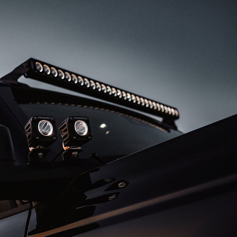 Rigid Revolve 20" Bar installed on a vehicle: A low-angle view of a dark vehicle's roofline with a Revolve light bar above the windshield and two Rigid pods at the A-pillar