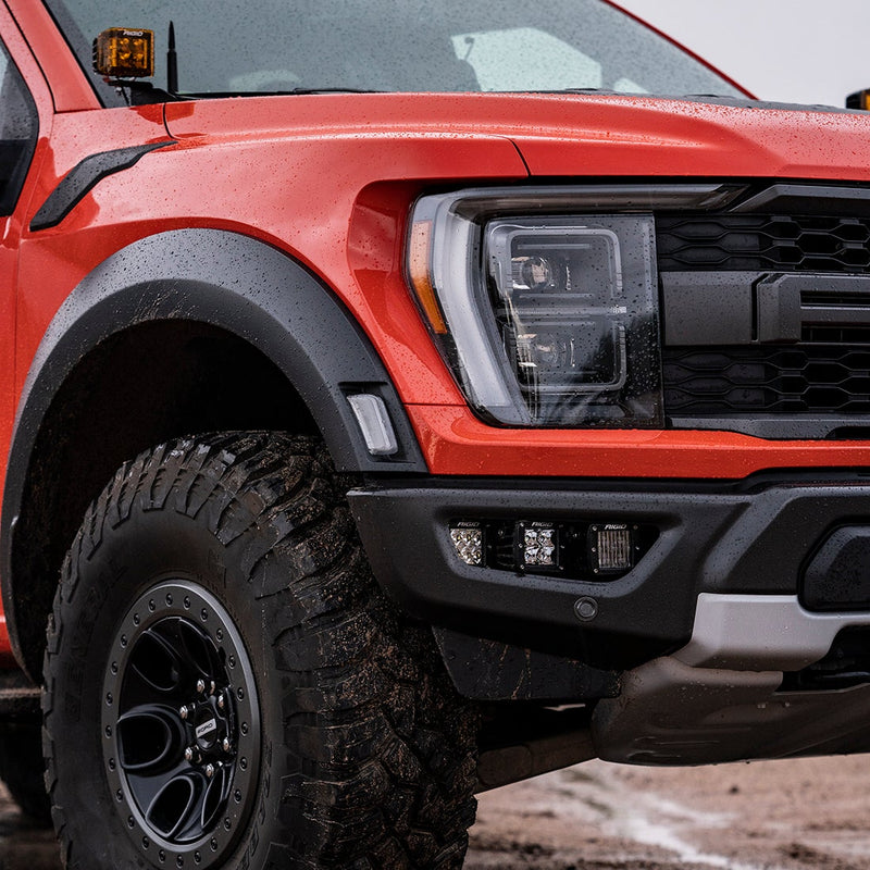 Rigid Triple Fog Light Kit installed on a Ford F-150 Raptor: front corner of a red Ford F-150 Raptor with square pods in the bumper opening and an amber pod at the A-pillar