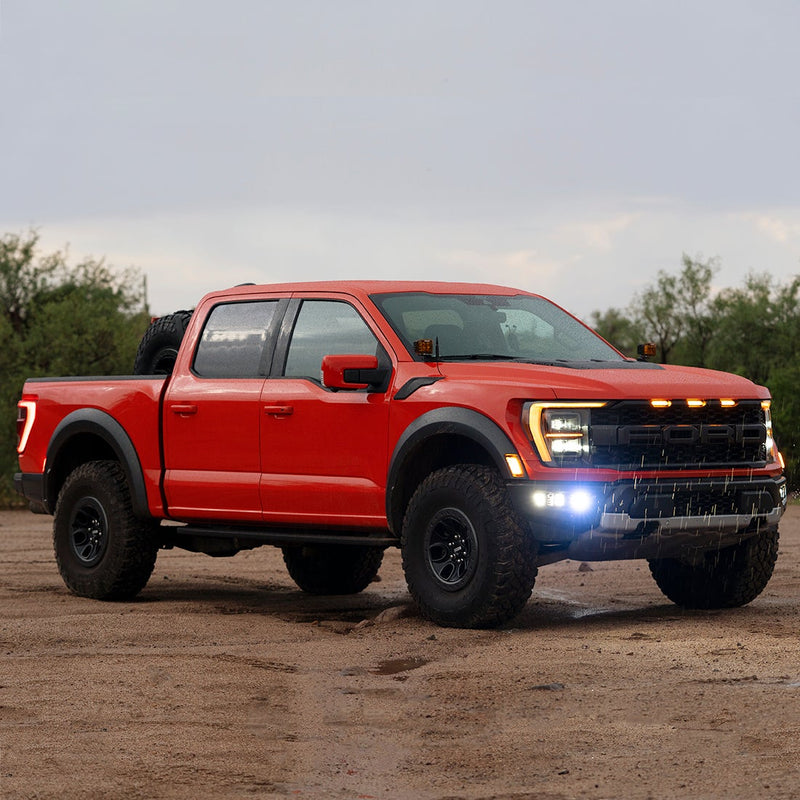 Rigid Triple Fog Light Kit installed on a Ford F-150 Raptor: red Ford F-150 Raptor on wet sand with lit fog pods in the bumper and amber A-pillar pods