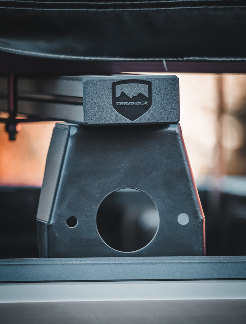 Close-up of the TeraFlex Cross Bar Kit: close-up of a black TeraFlex shield-logo riser block on a bed rail with a tent bag above