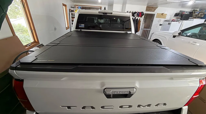 Worksport NEXUS hard folding tonneau cover on a pickup: customer photo from behind a white Toyota Tacoma in a garage with a closed black hard trifold cover
