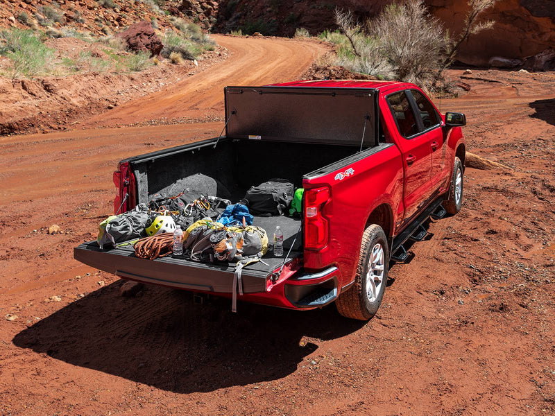 UnderCover ArmorFlex hard folding tonneau cover folded fully open against the bulkhead with the tailgate down and the bed loaded with outdoor gear, high rear three-quarter view on a desert track.