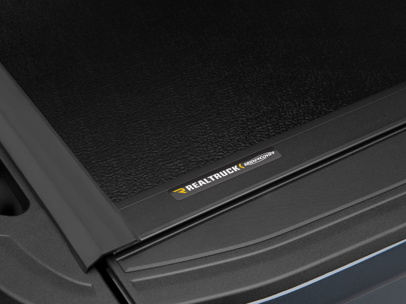 Close-up of the RealTruck UnderCover nameplate badge on the textured gloss-black panel surface of the Flex cover.