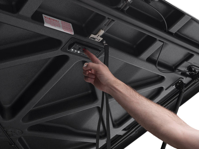 A hand pressing the button on the LED light module set into the black textured underside of the raised Elite lid.