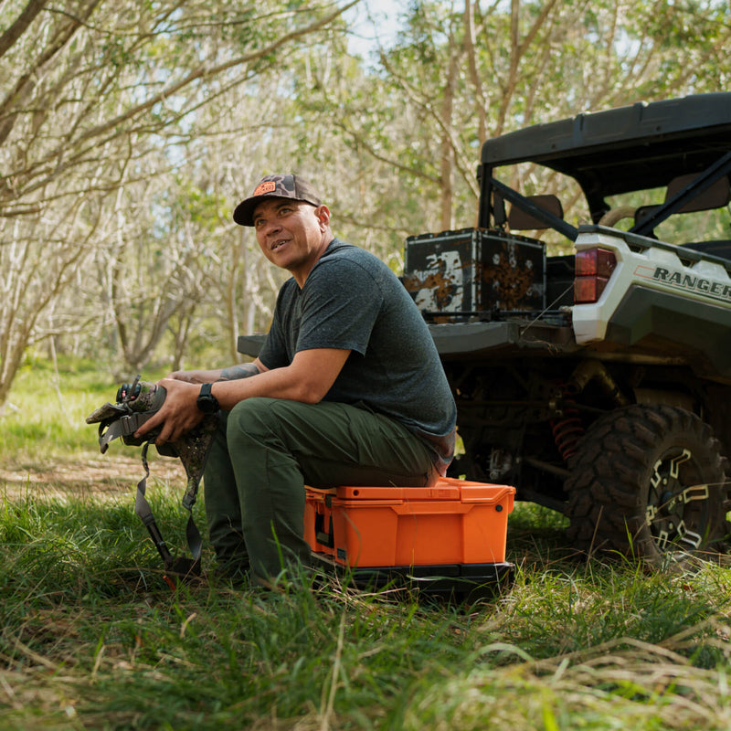Hunter sitting on a closed Blaze Orange DECKED Halfrack 32 beside a side-by-side in tall grass