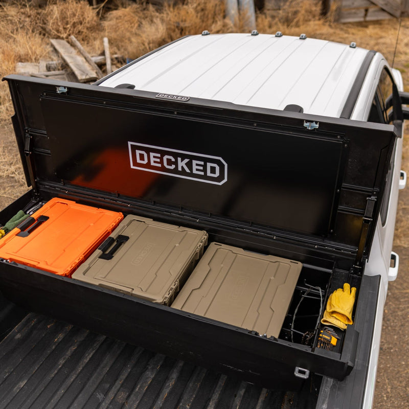 Two Desert Tan DECKED Halfrack 32 cases resting on the interior tray of an open black DECKED Tool Box in a pickup bed, next to a Blaze Orange case