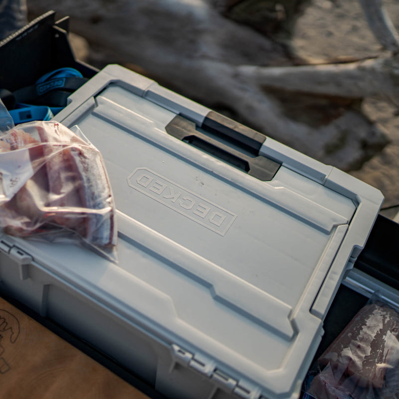 Cool Gray DECKED Halfrack 32 closed on a tailgate beside driftwood, with bagged fillets resting on the lid and a black DECKED case of straps behind