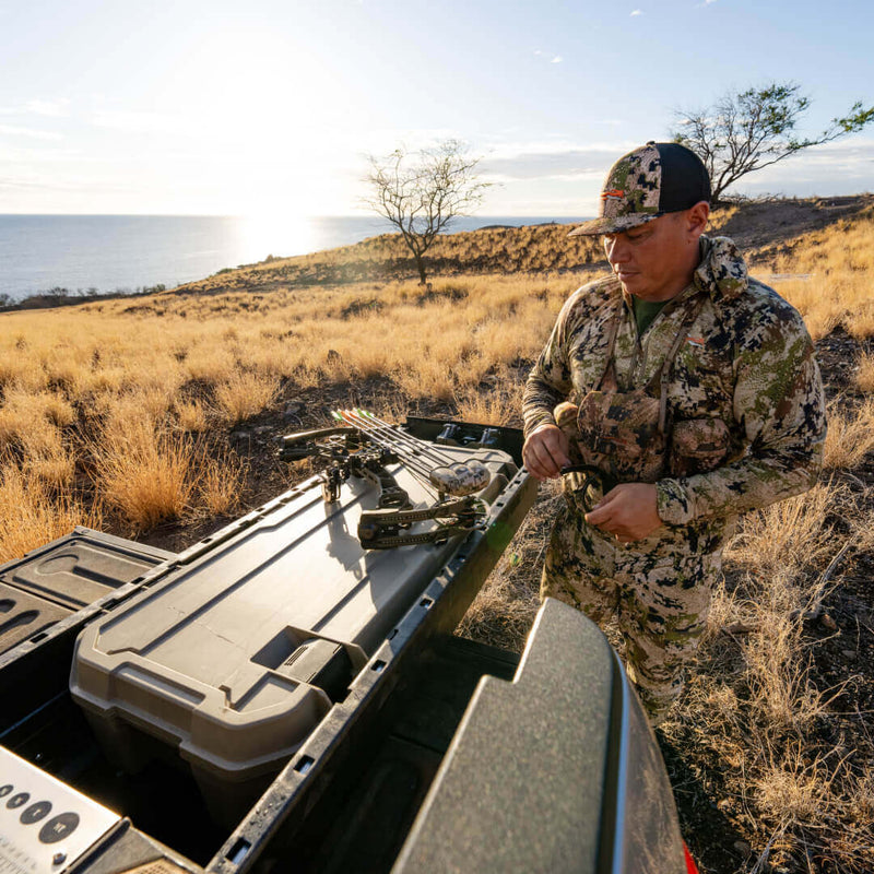 Hunter resting a compound bow on the closed lid of a Desert Tan DECKED Honcho 80 in a truck bed at sunset