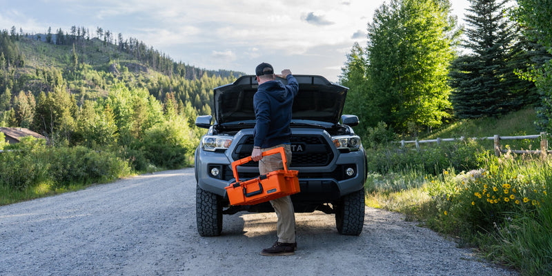 Driver carrying the Blaze Orange DECKED Roadside Emergency OS Kit to the raised hood of a pickup on a mountain road