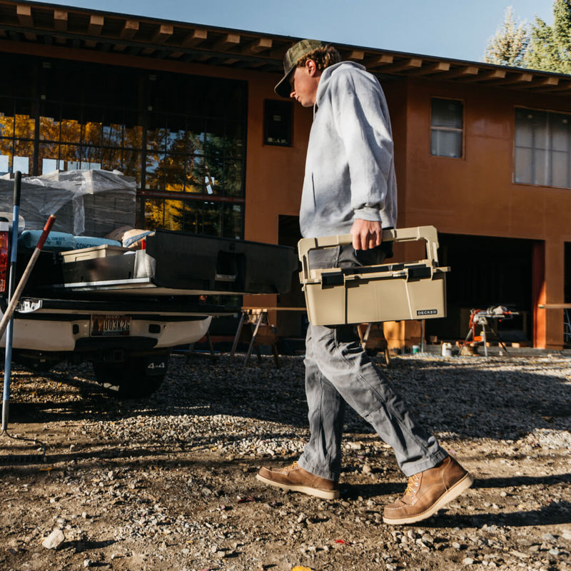 Worker carrying the Desert Tan DECKED Sixer 16 by its lid handle across a jobsite