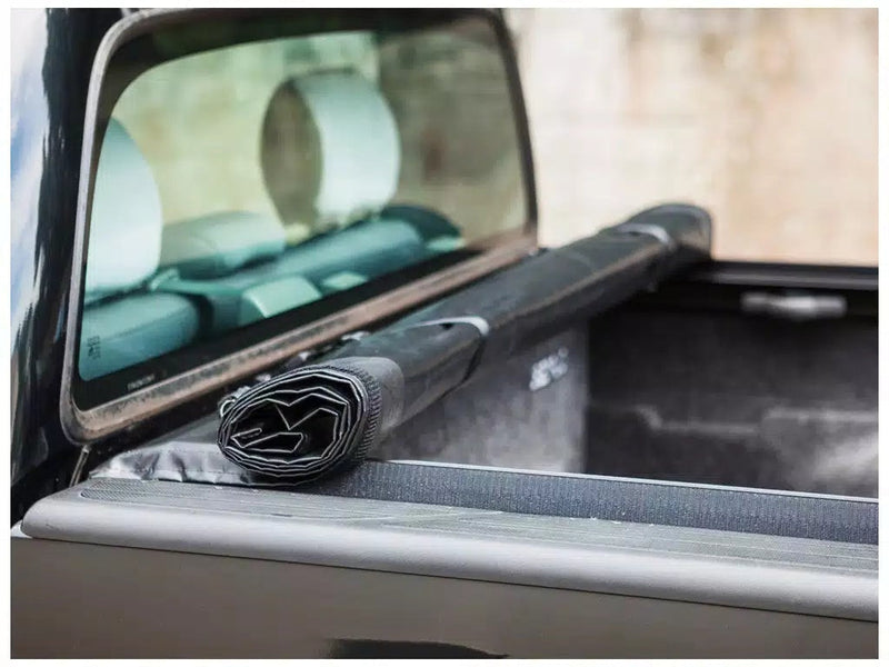Close-up of the Access Limited Edition roll-up tonneau cover: rolled vinyl cover strapped at the cab window on a side rail, carpeted bed liner beyond (bedliner shown, not included)