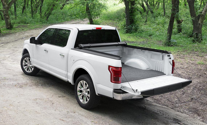 Access Limited Edition roll-up tonneau cover on a pickup bed: white crew cab pickup on a forest road with the roll-up cover rolled to the cab, tailgate down, bed mat (bed mat shown, not included)