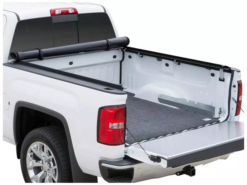 Full view of the Access Limited Edition roll-up tonneau cover: white pickup on white with the roll-up cover rolled to the cab, tailgate down, carpet mat in bed (bed mat and trailer hitch receiver shown, not included)