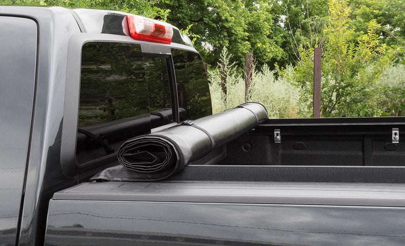 Close-up of the Access LiteRider roll-up tonneau cover: black vinyl cover rolled up and strapped behind the cab of a gray pickup, bed open (bedliner shown, not included)