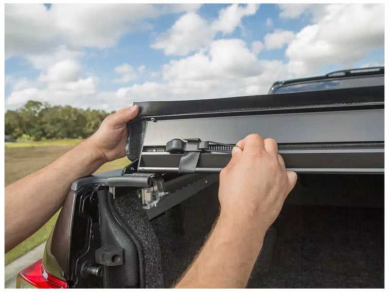 Close-up of the Access LiteRider roll-up tonneau cover: hands lifting the rear edge of a roll-up cover to show the tension bar and latch at the tailgate (bed mat shown, not included)