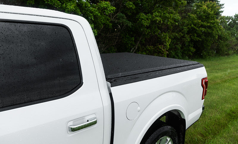 Access Lorado roll-up tonneau cover on a pickup bed: rain-beaded black roll-up cover closed flat over the bed of a white crew-cab pickup, side view from the cab