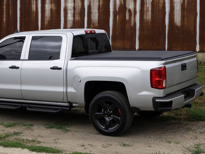 Black vinyl roll-up tonneau cover closed flat over a pickup bed, rear three-quarter view by a rusted metal wall