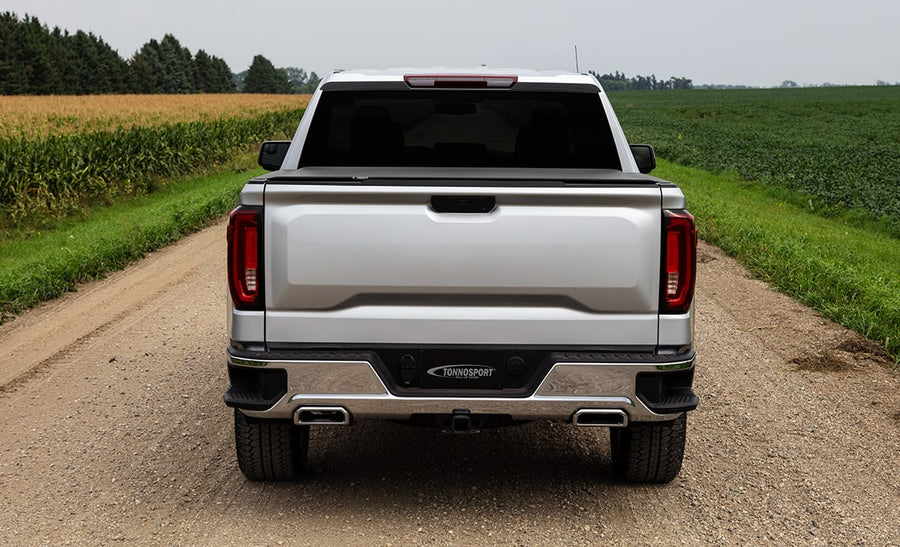 Access TonnoSport roll-up tonneau cover: Rear view of a silver pickup on a gravel farm road, low-profile roll-up cover closed over the bed