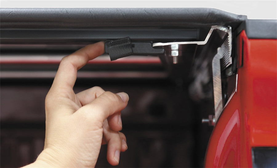 Access TonnoSport roll-up tonneau cover: Finger pulling a latch release under the rear edge of a roll-up cover on a red pickup