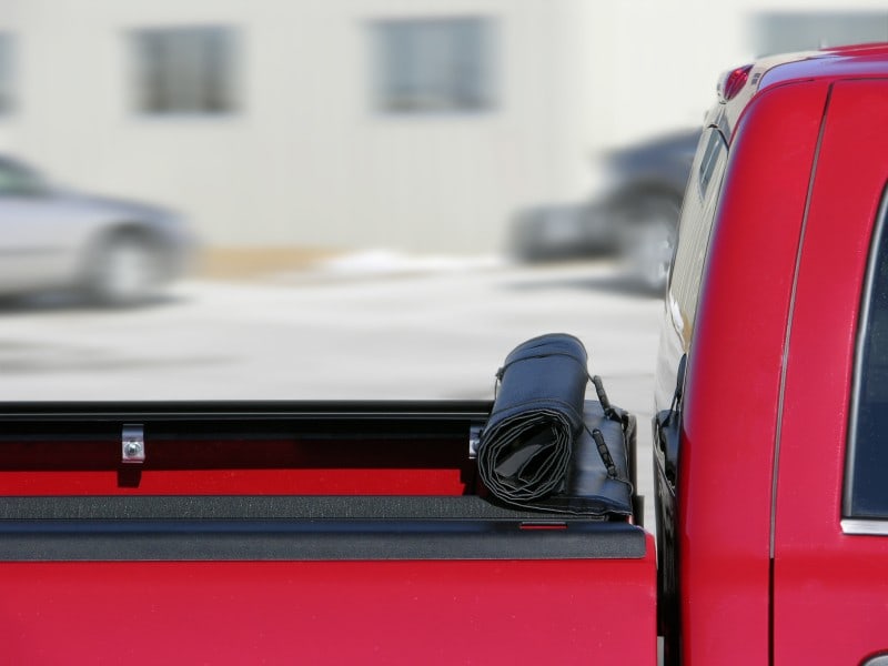 Access Lorado roll-up tonneau cover: Black cover rolled up and strapped at the front of a red pickup bed, side rail visible