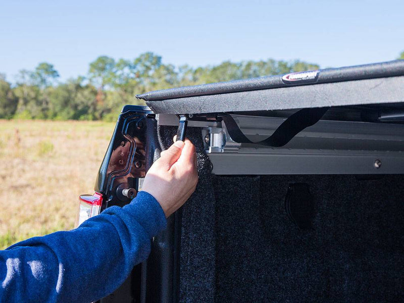 Hand pulling the latch at the rear corner of the vinyl roll-up cover, tailgate open, carpeted bed