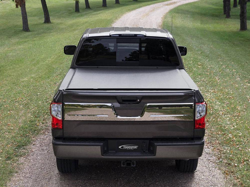 Straight rear view of a dark pickup on a gravel lane, low-profile roll-up cover closed over the bed