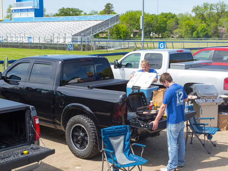 Two men tailgating beside a black pickup with its roll-up cover rolled forward, grill and cooler at the tailgate