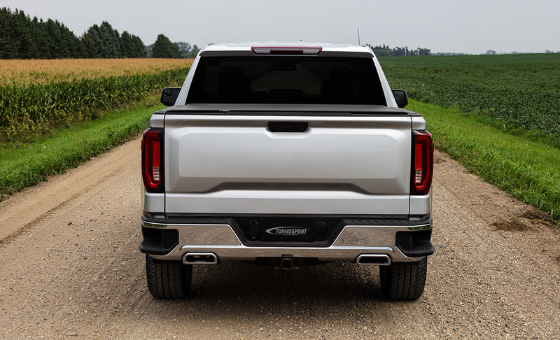 Access TonnoSport roll-up tonneau cover on a pickup bed: rear view of a silver pickup on a gravel farm road, low-profile roll-up cover closed over the bed