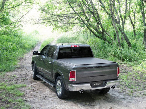 Rear three-quarter view of a black soft roll-up cover closed flat over a pickup bed, parked on a wooded dirt track, Vanish logo plate