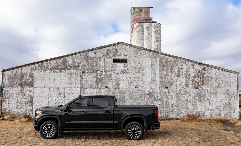 Access Vanish roll-up tonneau cover on a pickup bed: black crew cab pickup parked in profile before a weathered barn and silo, cover closed flush with bed rails (running boards shown, not included)