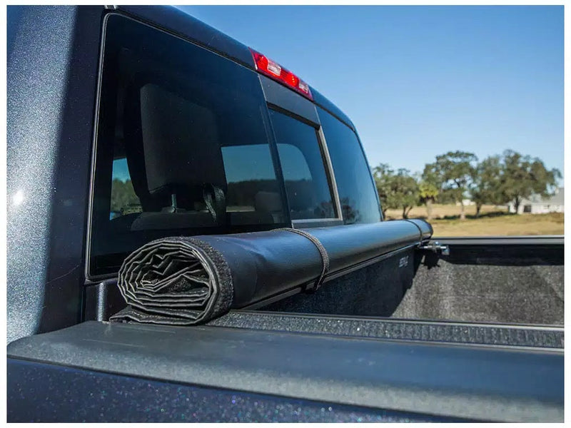 Close-up of the Access Vanish roll-up tonneau cover: close-up of a black vinyl cover rolled and strapped against the cab end of a pickup bed (carpeted bed liner shown, not included)