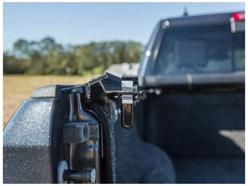 Close-up of the Access Vanish roll-up tonneau cover: close-up of an aluminum side rail and clamp at the rear corner of a pickup bed, cab blurred behind (carpeted bed liner shown, not included)