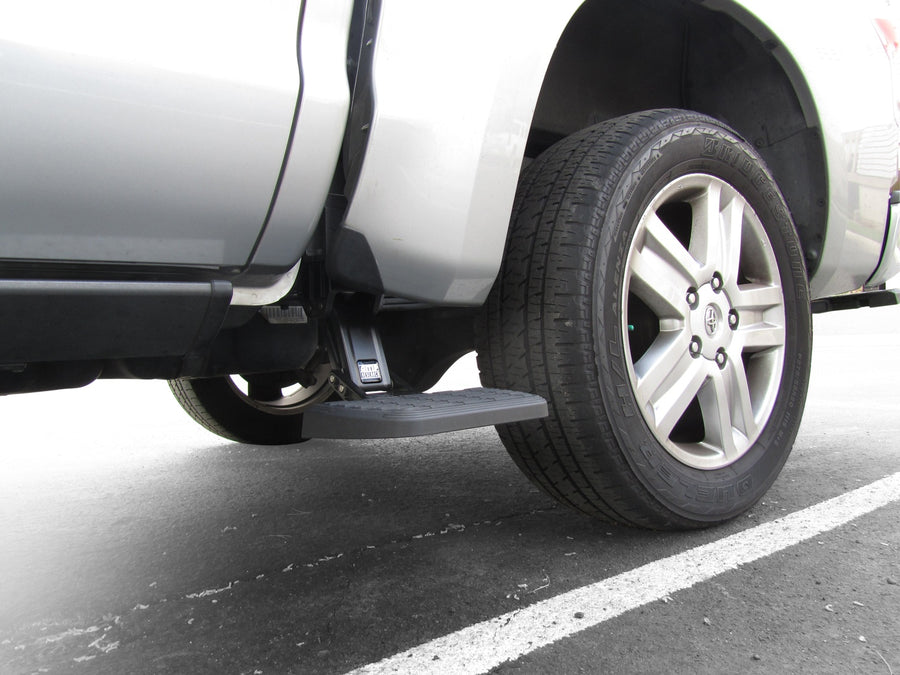 Side bed step deployed ahead of the rear wheel of a silver pickup in a parking lot
