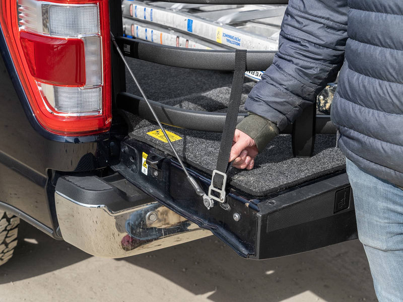 A hand tightens the BedXTender HD Sport strap at the lowered tailgate beside the tail lamp of a dark blue pickup