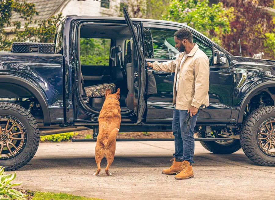A man opens the rear door of a lifted dark blue pickup as his dog jumps up toward the cab, running board below
