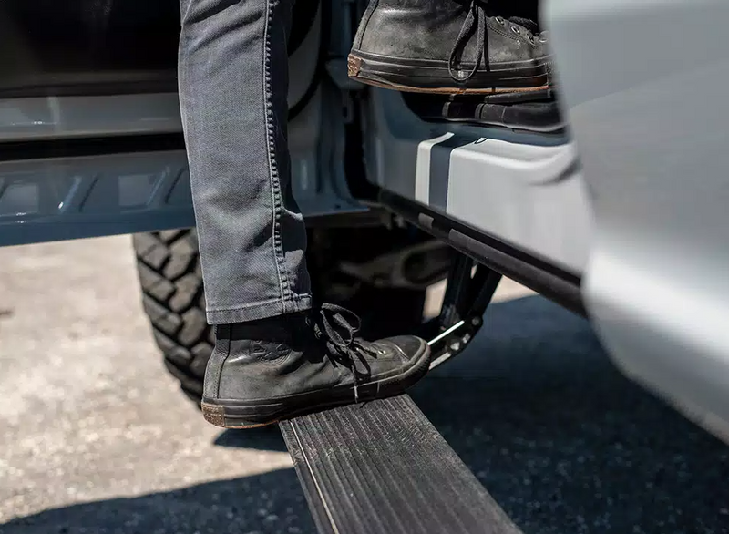 A person in jeans and sneakers steps up from the deployed running board into a silver pickup