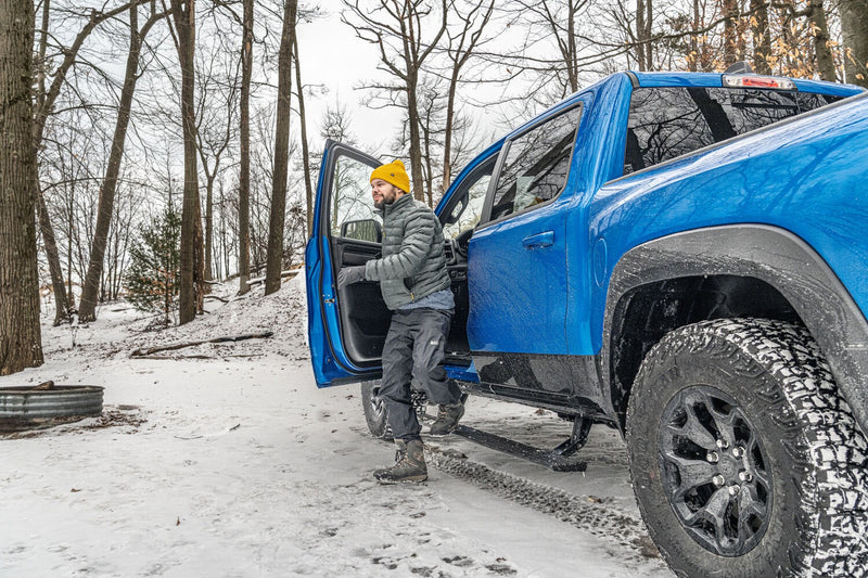 Man in a yellow beanie climbing out of a blue pickup onto the deployed running board in a snowy forest.