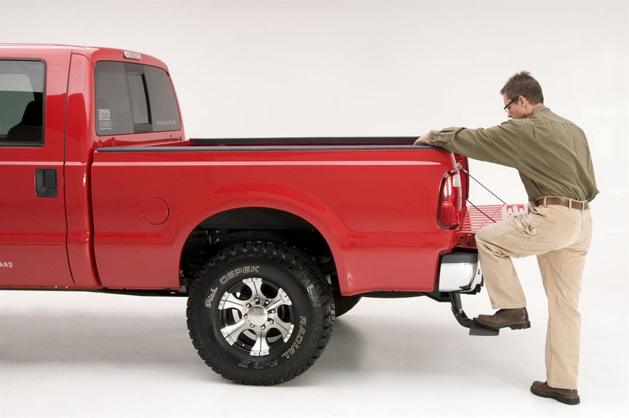 A man stands on the BedStep to reach over the bed side of a red heavy-duty pickup, studio background