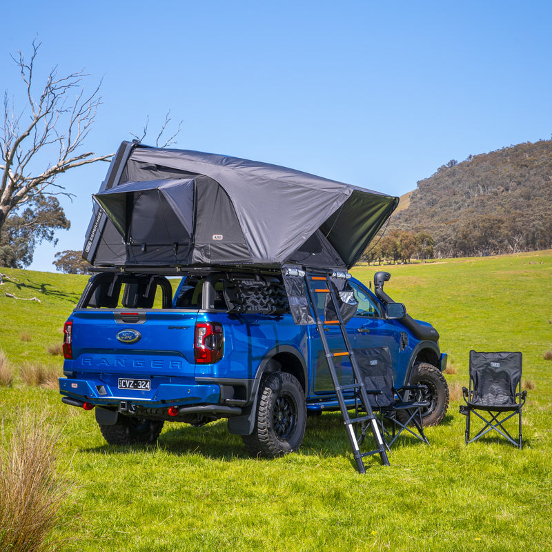 ARB Esperance V2 Roof Top Tent 802200V2 deployed on the roof rack of a blue Ford Ranger in a grass paddock, ladder extended with two ARB camp chairs alongside