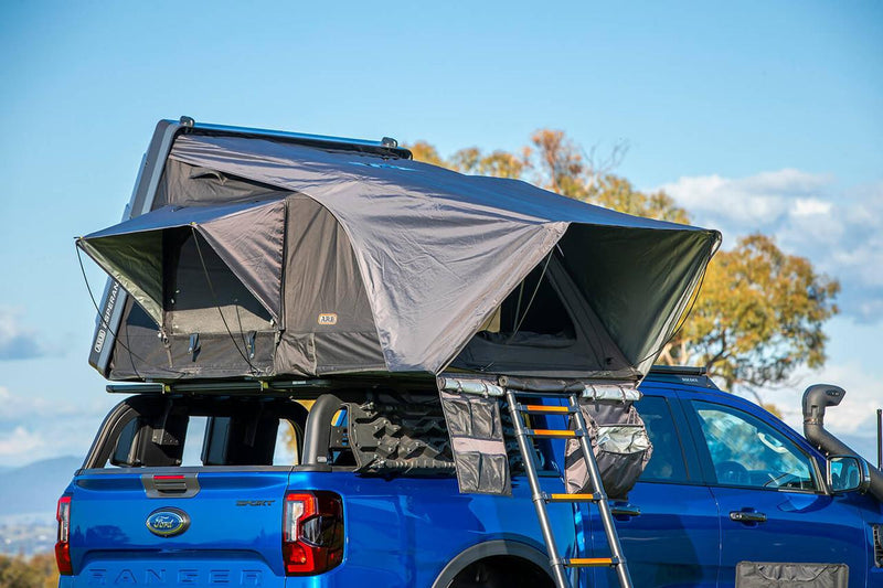 Close lifestyle view of the ARB Esperance V2 Roof Top Tent 802200V2 pitched on a blue Ford Ranger roof rack, showing rainfly, window awnings and extended ladder