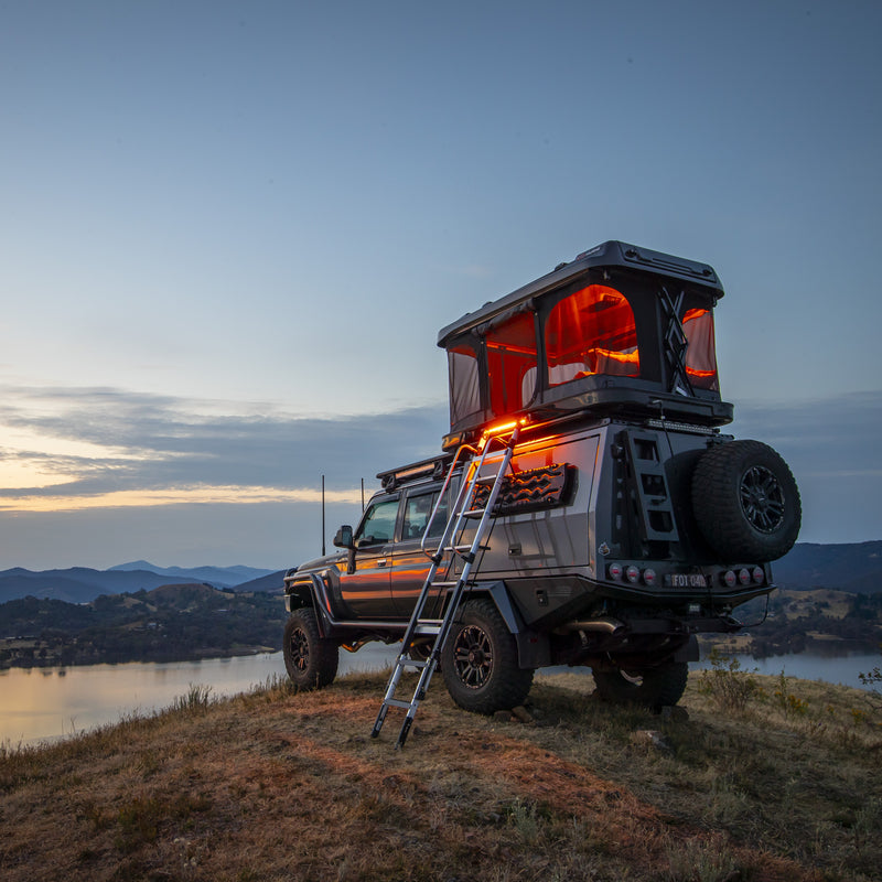 ARB 802500 Altitude Rooftop Tent open and lit at dusk on a Toyota 79-Series tray truck overlooking a lake, ladder deployed to the ground