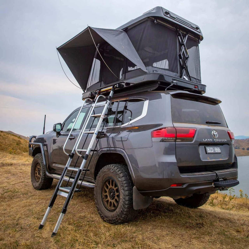 ARB 802500 Altitude Rooftop Tent deployed on a Toyota Land Cruiser 300 with awning flap propped and ladder extended on a grassy hillside
