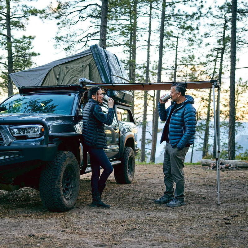 ARB 814301 soft case awning deployed in sand canvas off a Toyota pickup in a pine forest with two campers standing beneath it